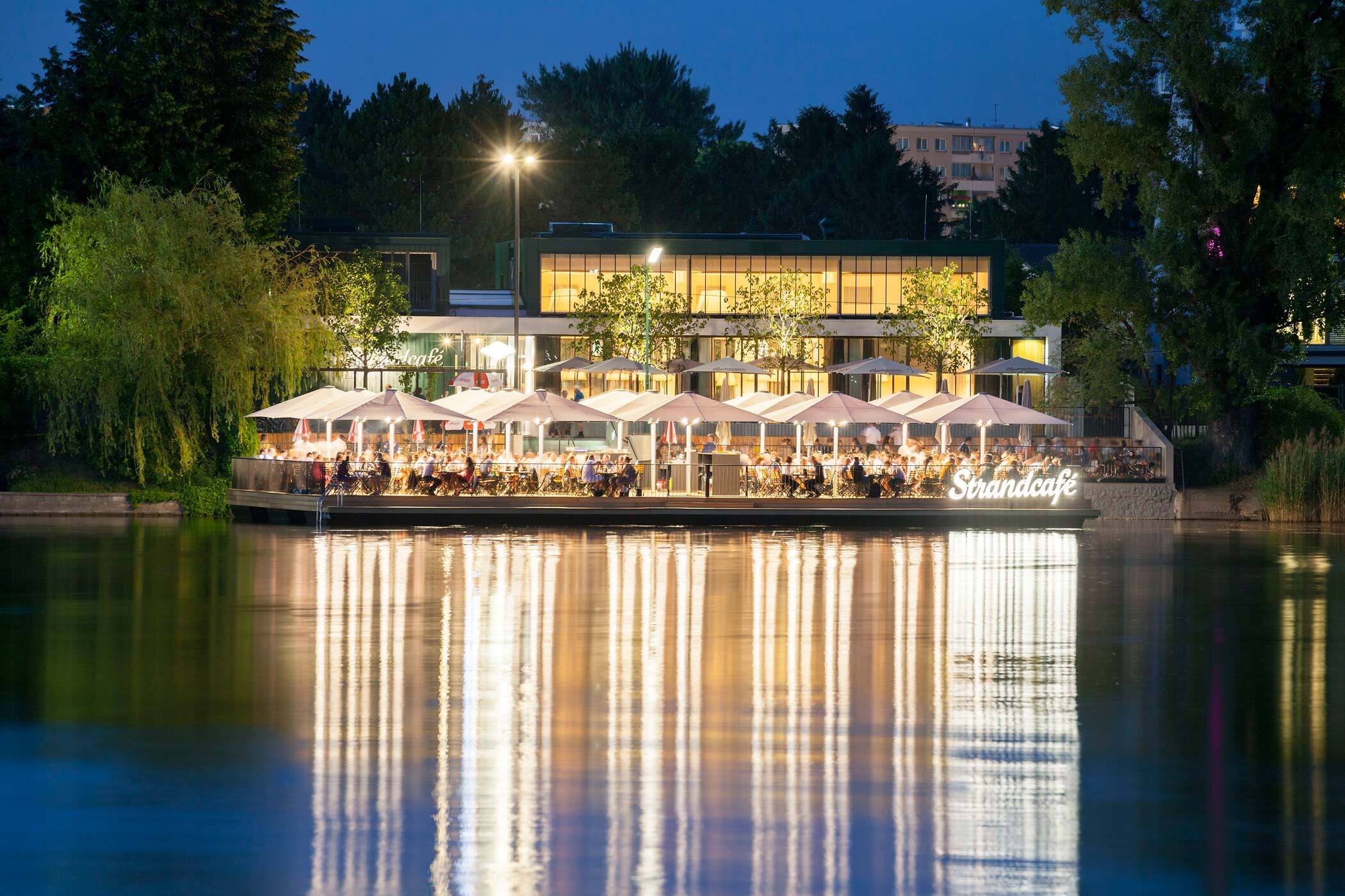 Strandcafe auf einer Wasserplattform bei Nacht, Gäste speisen unter Sonnenschirmen; beleuchtete Fassade und Spiegelungen im Wasser.