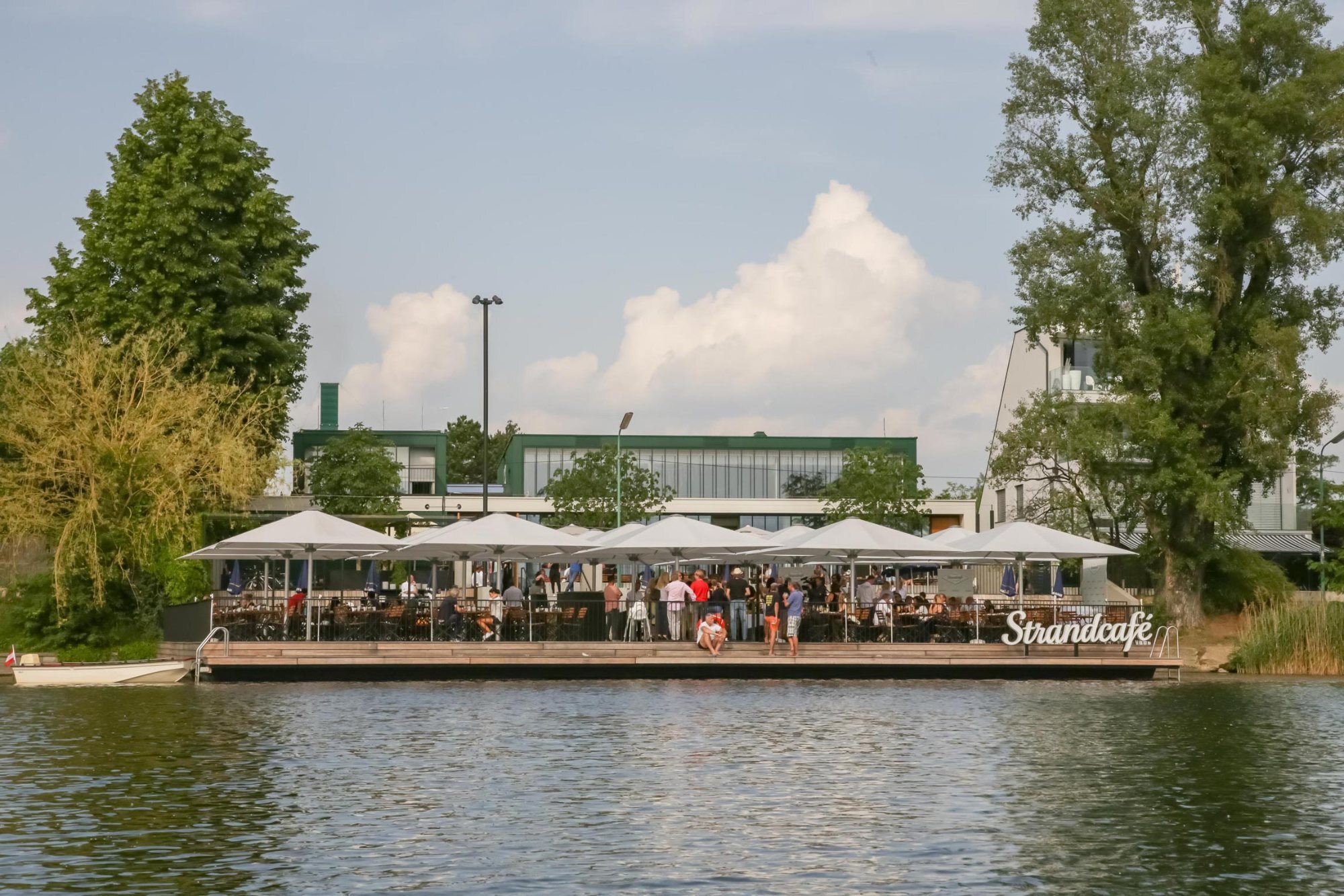 Freiluftcafé auf dem Pier am Wasser; Gäste sitzen unter weißen Sonnenschirmen und speisen; Hintergrund Gebäude und Bäume, Schild Standcafe sichtbar.